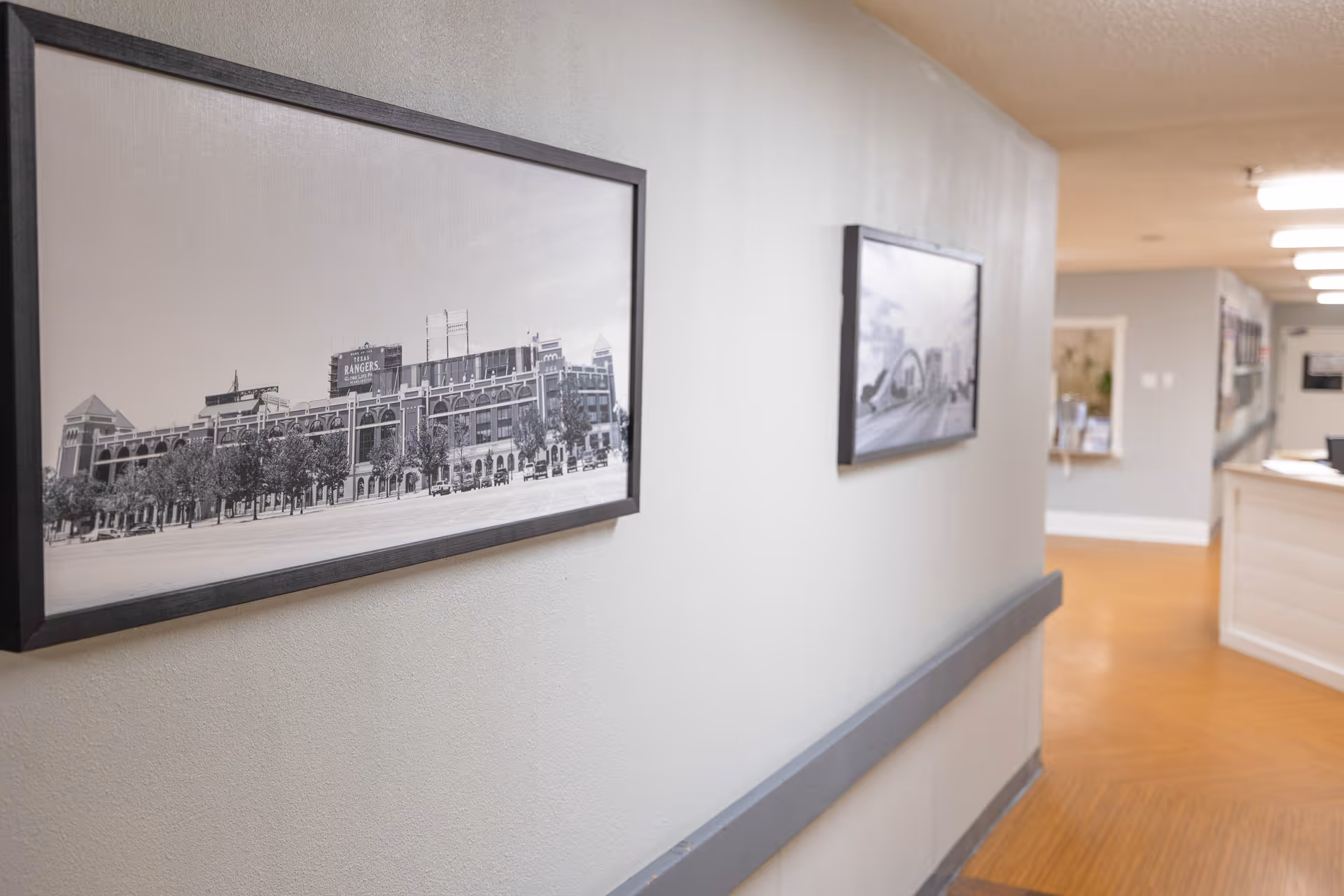 A hallway in a nursing and rehab facility with framed black and white photographs on the wall. The floor has a wood-like finish, and there is a handrail along the wall. The hallway leads to a reception or nurse's station area with bright lighting.