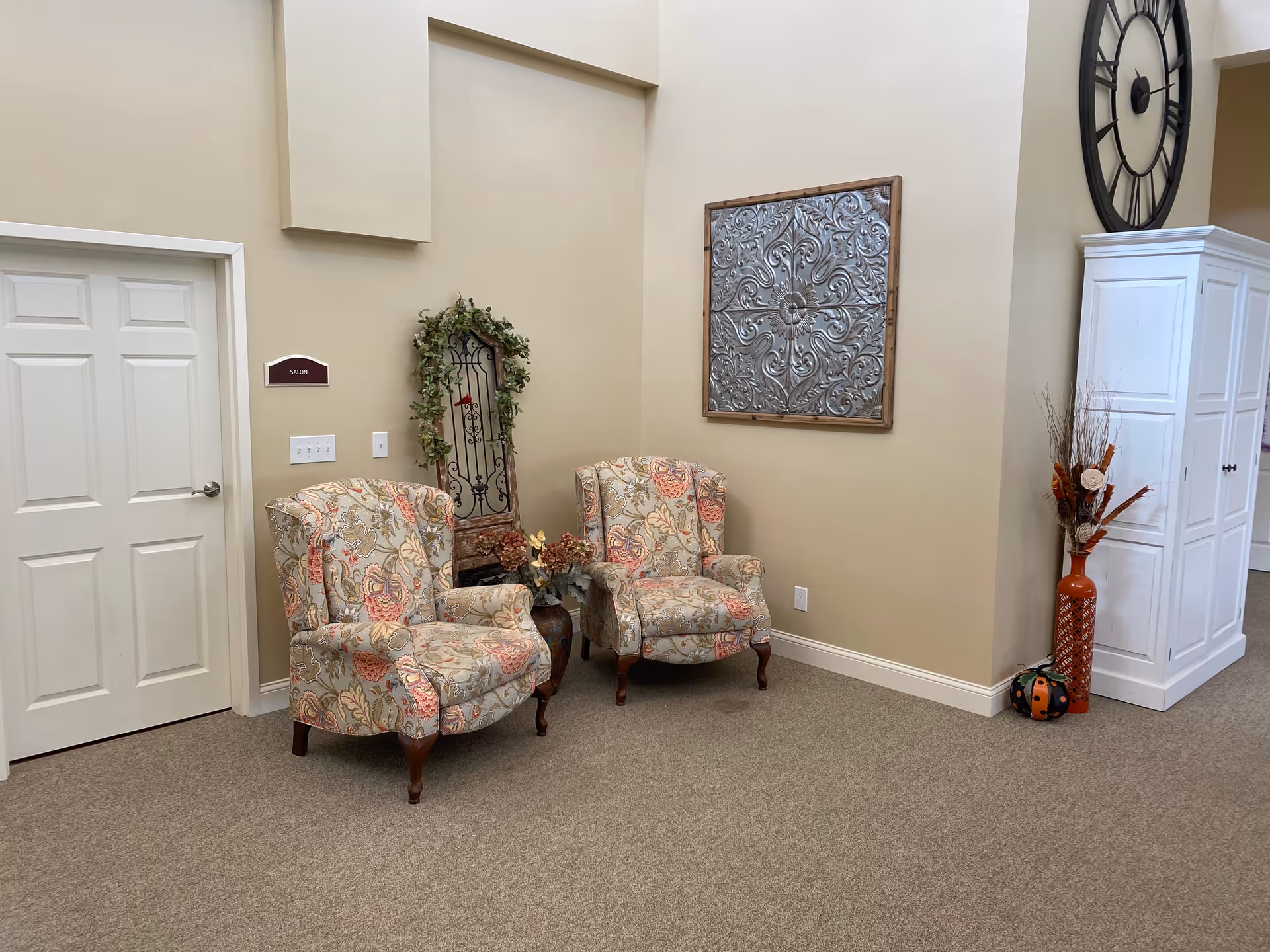 Two patterned armchairs in a decorated seating area with wall art, a large clock, and a white storage cabinet in a senior living facility.
