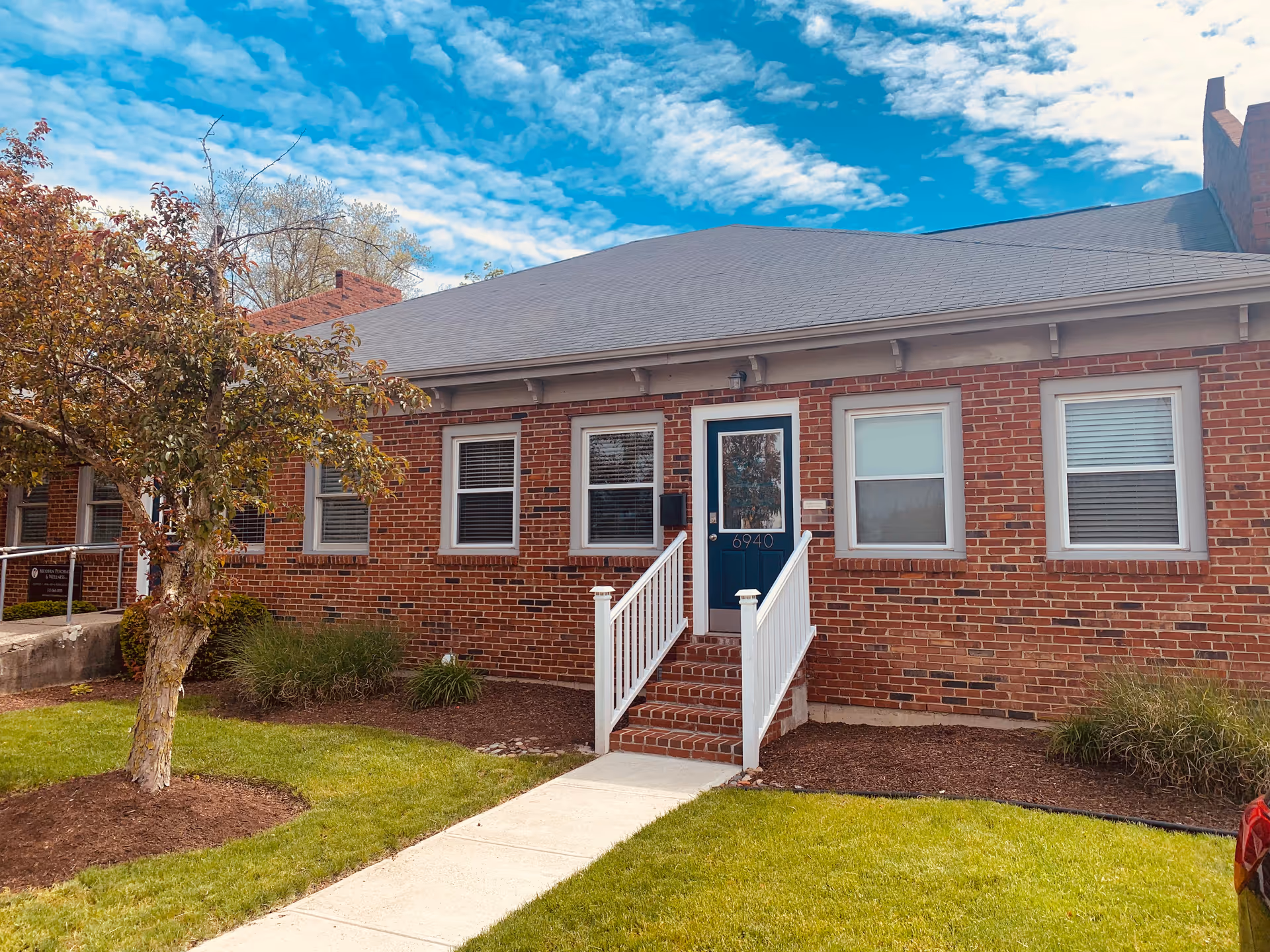 Brick senior living building front with white stairs, blue door, windows, and landscaped yard.