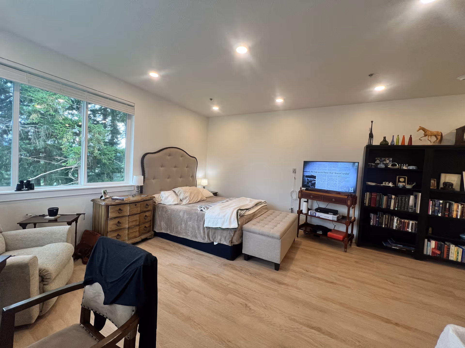 Cozy memory care bedroom featuring a tufted headboard, dresser, seating area, and a bookshelf.