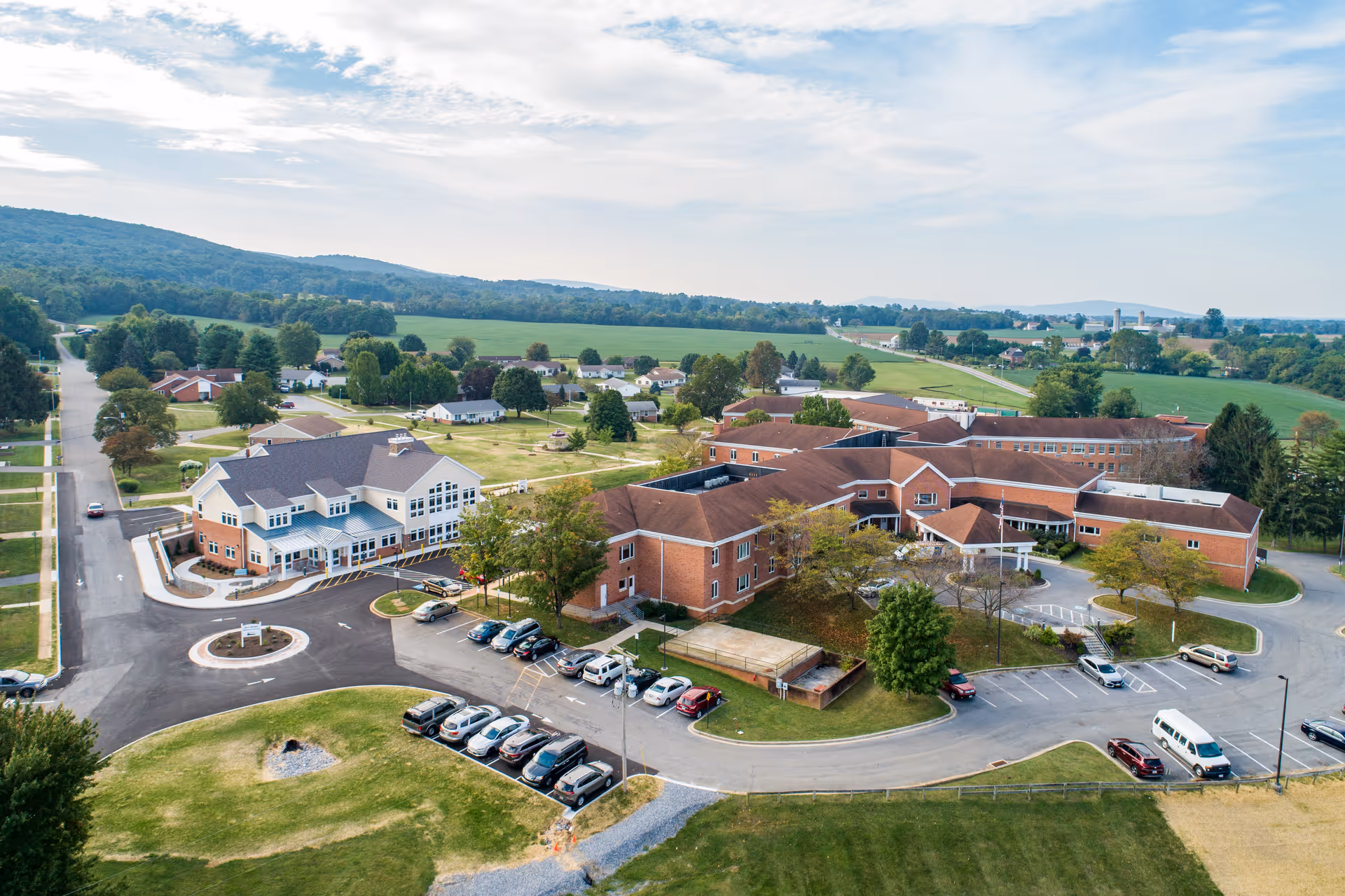 Aerial view of Fahrney-Keedy Senior Living Community showing multiple connected brick buildings with red roofs, surrounded by parking lots, green lawns, trees, and a rural landscape with hills and fields in the background under a partly cloudy sky.