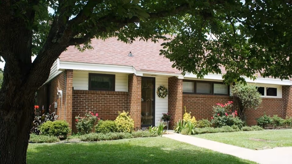 Exterior view of a single-story brick building with a sloped roof, surrounded by green grass, bushes, and flowering plants. A large tree with green leaves partially shades the front of the building, which has a glass door decorated with a wreath.