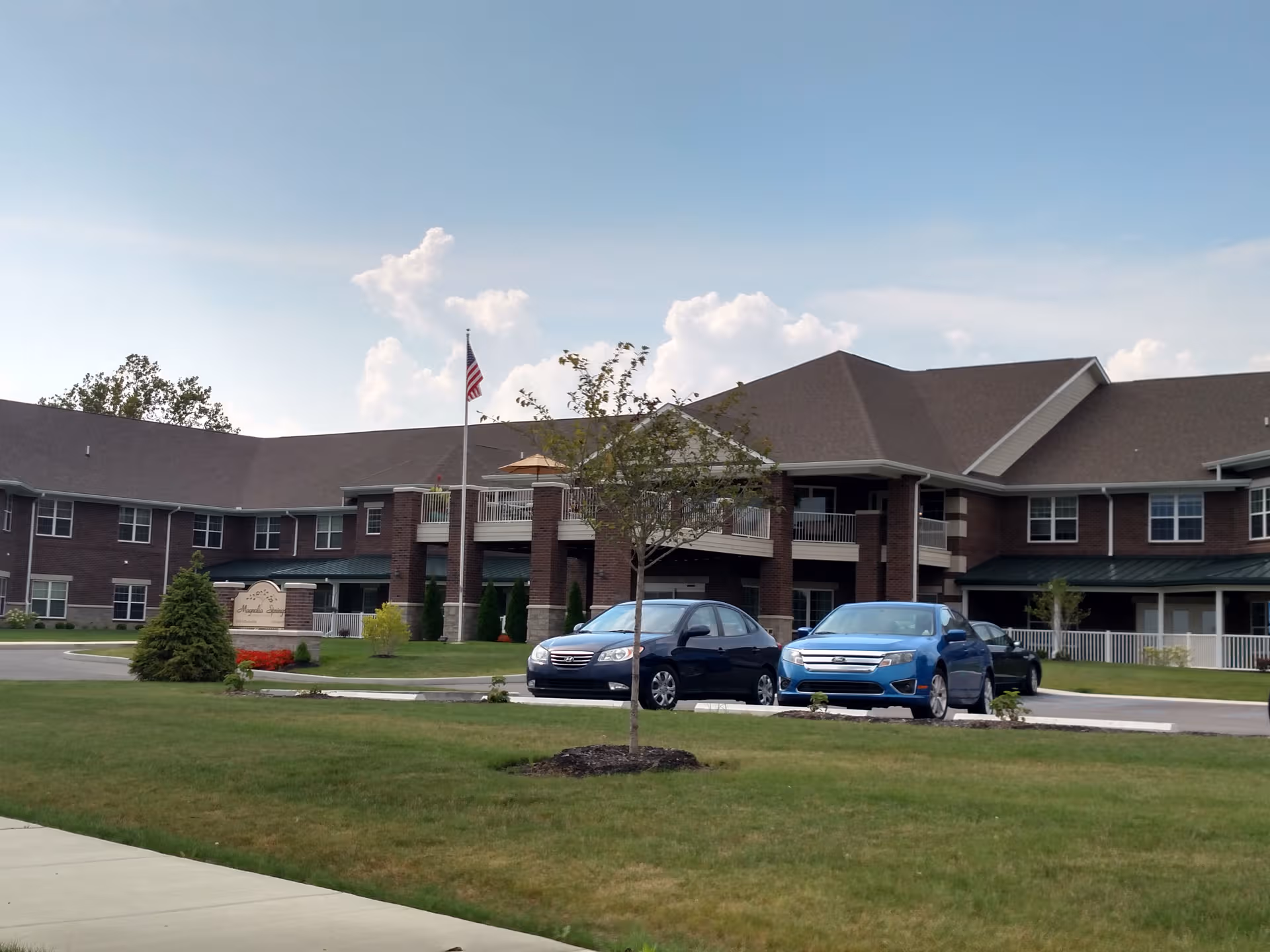 Exterior view of Magnolia Springs Southpointe, brick building with entrance, flagpole, and parked cars.
