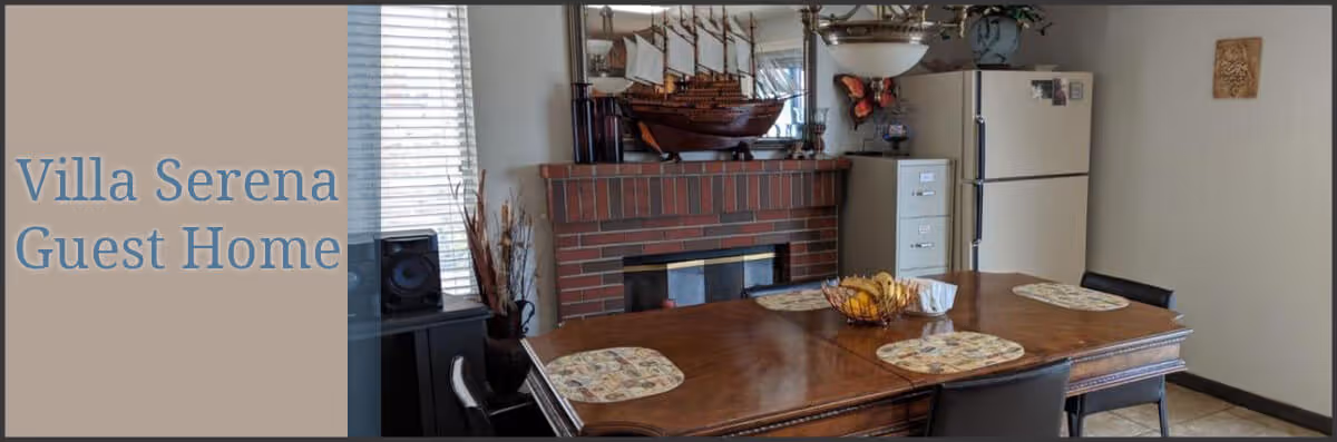 Dining area with a wooden table set with four placemats and a fruit bowl in the center. Behind the table is a brick fireplace with a decorative ship model on the mantel, a window with blinds, a white refrigerator, and a filing cabinet. The room has neutral-colored walls and tiled flooring.