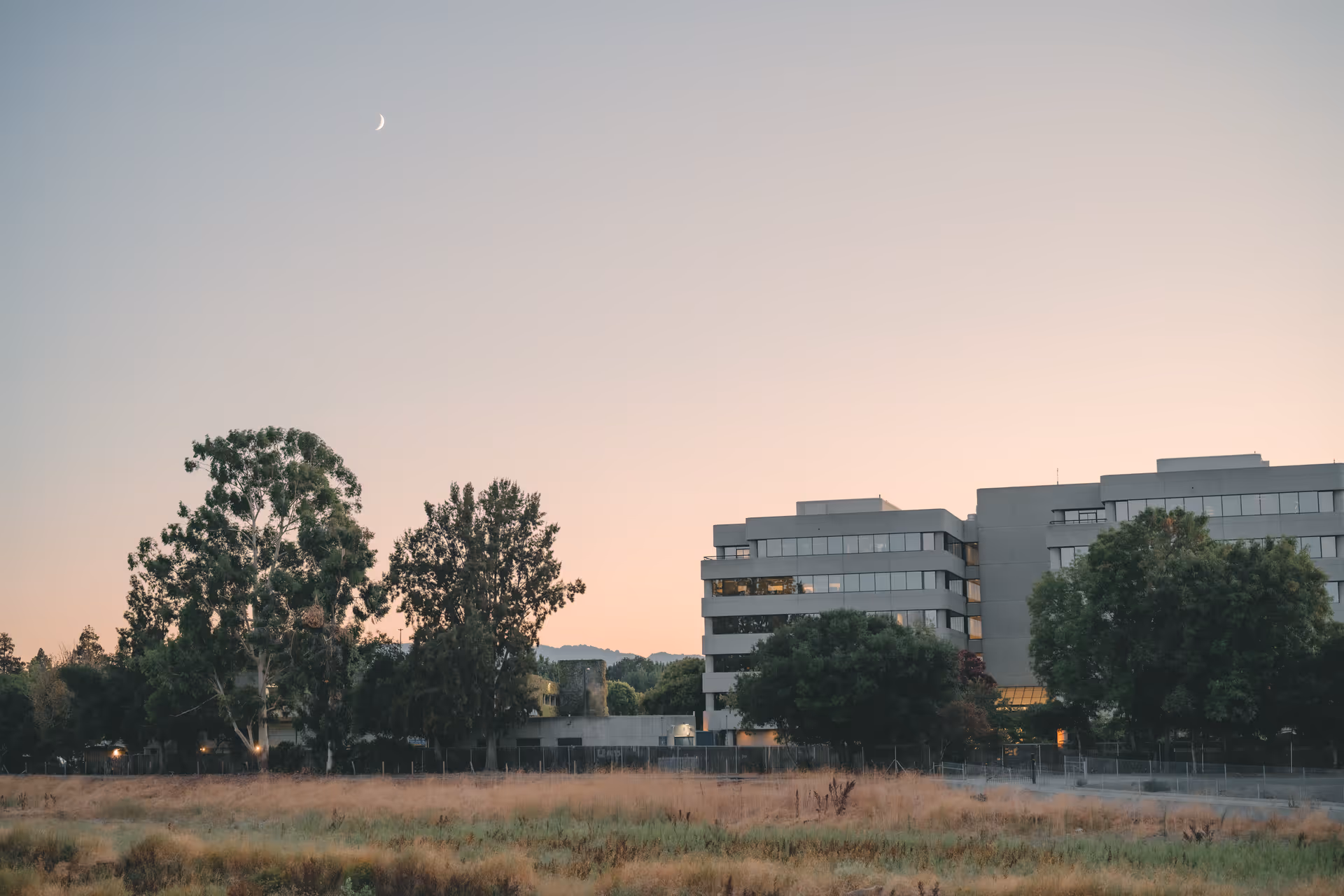 Sunset exterior view of a modern multi-story building with trees and open field.