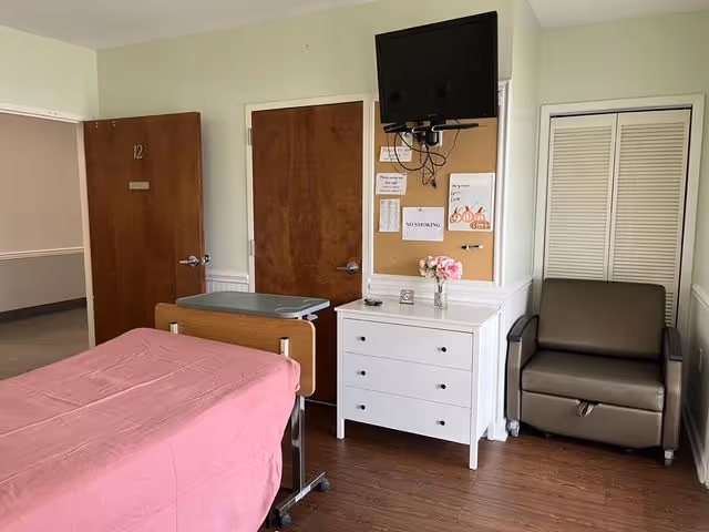 Abbey Hospice patient bedroom with bed, dresser, chair, mounted TV, and closet doors.