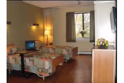 Shared patient bedroom with two floral beds, a small TV, window, and dresser.