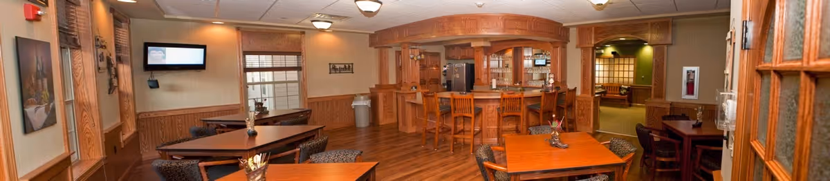 Interior view of a common area in a retirement community featuring wooden tables and chairs, a bar with high stools, wood-paneled walls, and warm lighting. There is a television mounted on the wall, decorative artwork, and a doorway leading to another room.