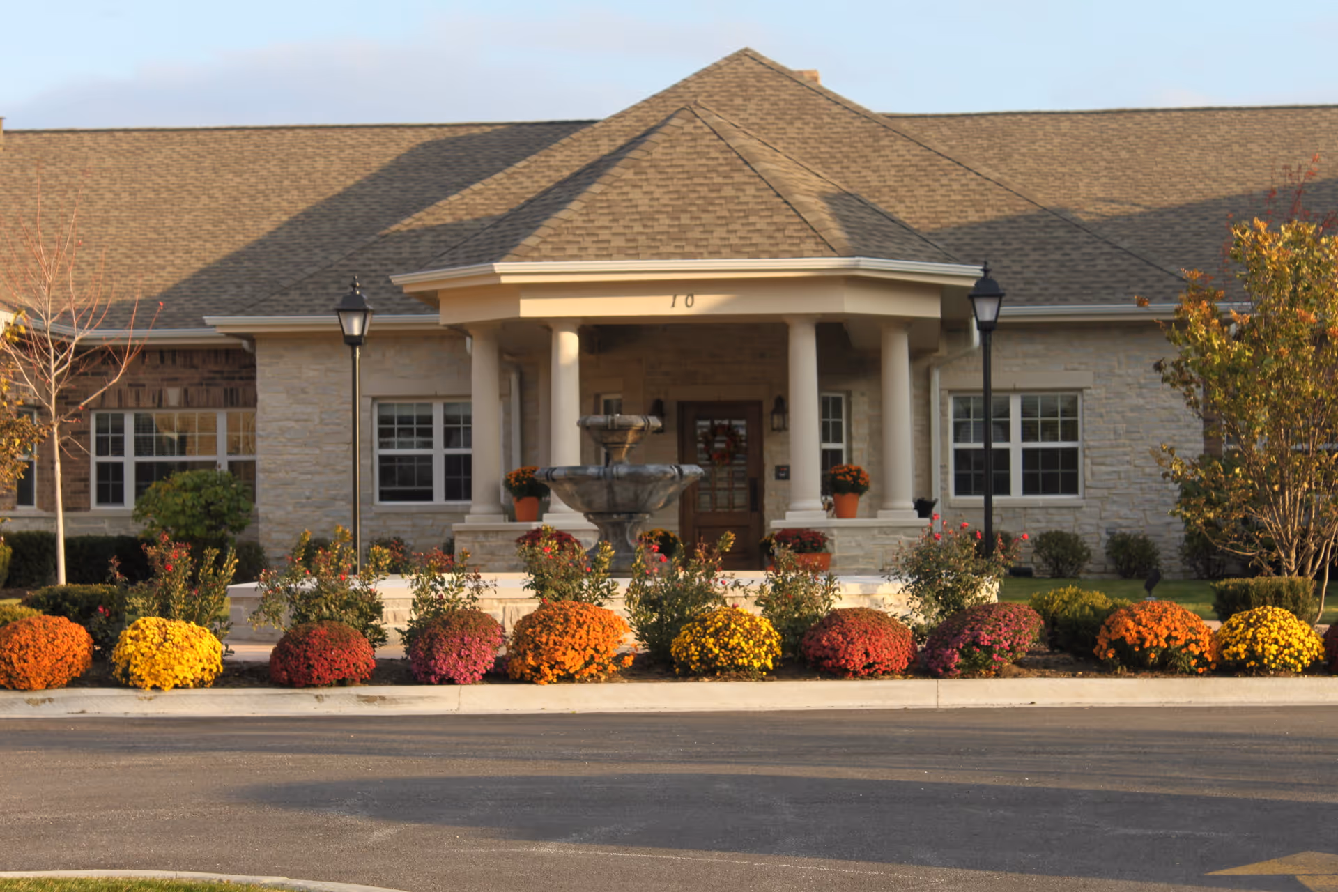 Front entrance of a senior living facility with columns, a central fountain, and colorful flowering shrubs.