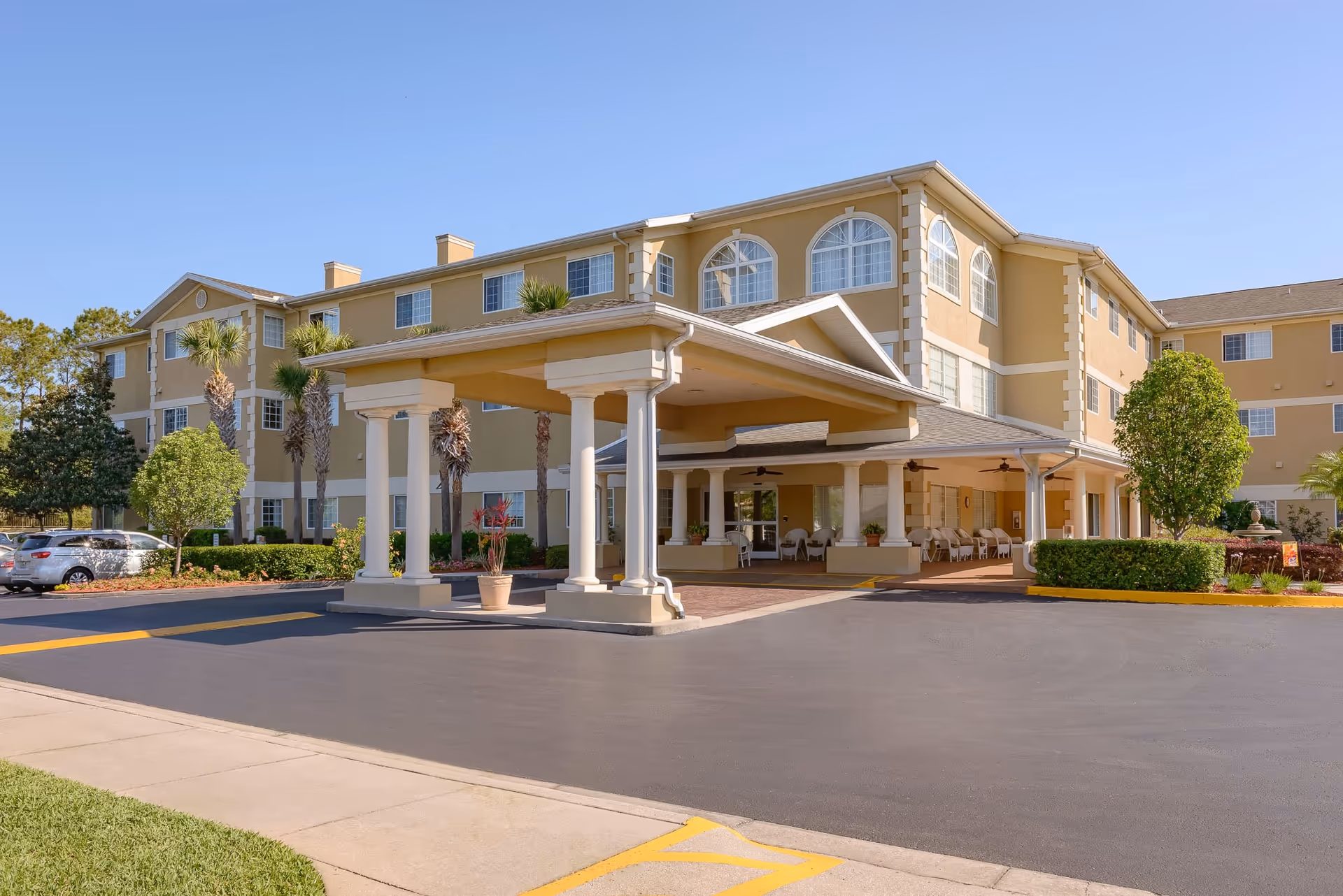 Exterior view of a three-story senior living facility building with a covered entrance supported by white columns, surrounded by palm trees, shrubs, and a parking area under a clear blue sky.