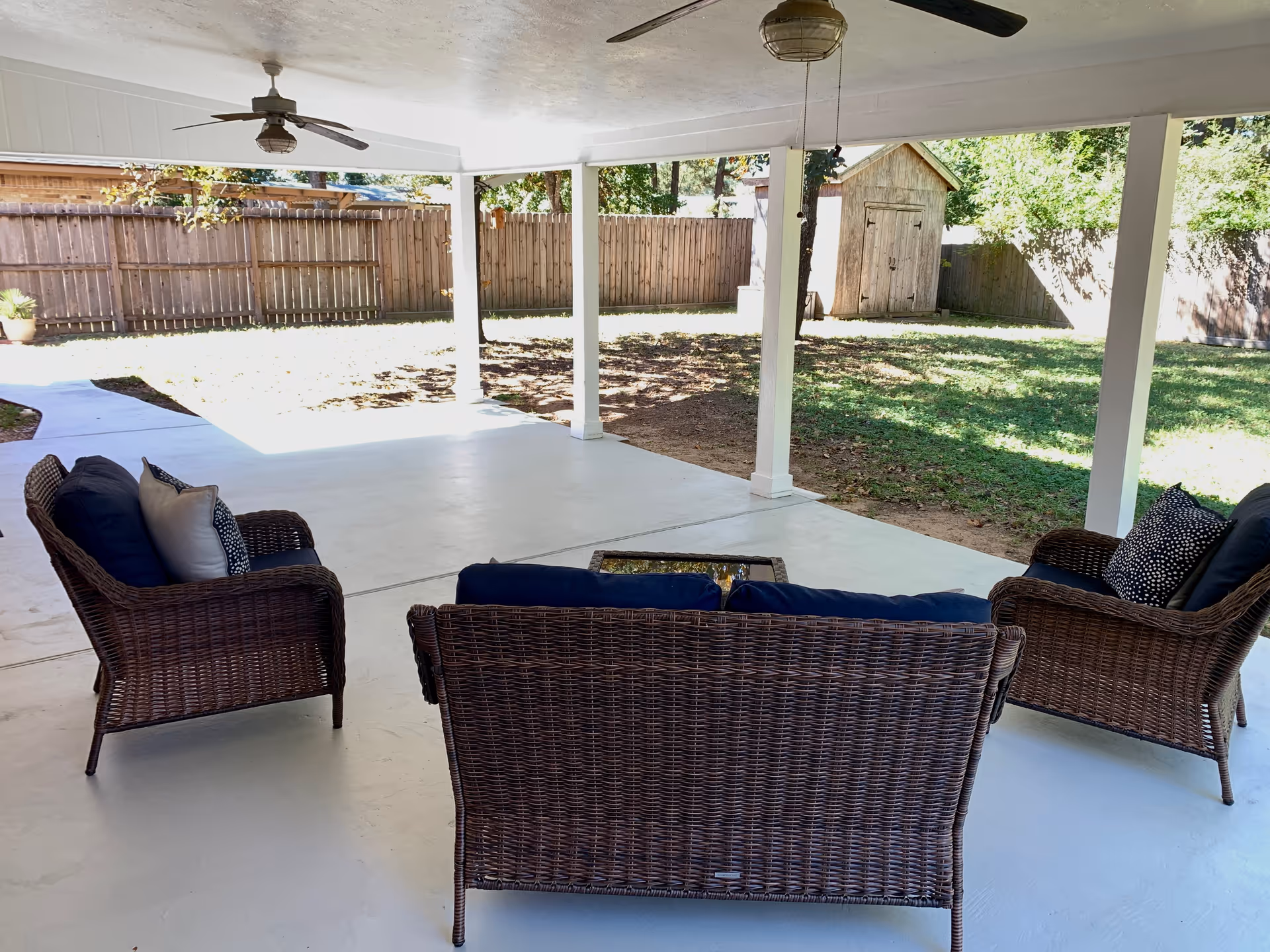 Covered outdoor patio with wicker seating and ceiling fans overlooking a fenced yard.