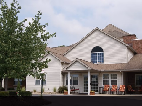 Exterior view of a senior living facility building with beige siding and a peaked roof. There is a covered entrance with a bench and several chairs with floral cushions arranged outside. A tree and some shrubs are visible in the landscaped area near the building.
