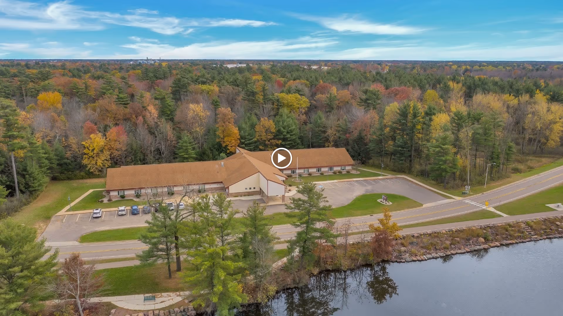 Aerial view of a single-story building with a brown roof surrounded by a parking lot, trees with autumn foliage, a road, and a body of water in the foreground.