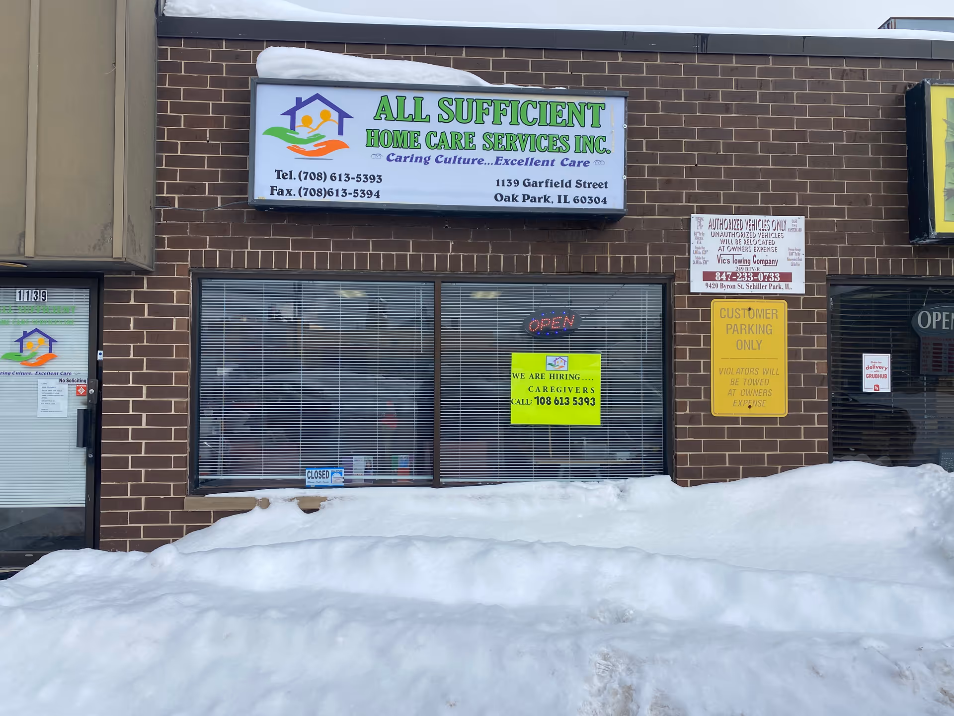 Brick storefront with All Sufficient Home Care Services sign, snow in foreground, windows with blinds.