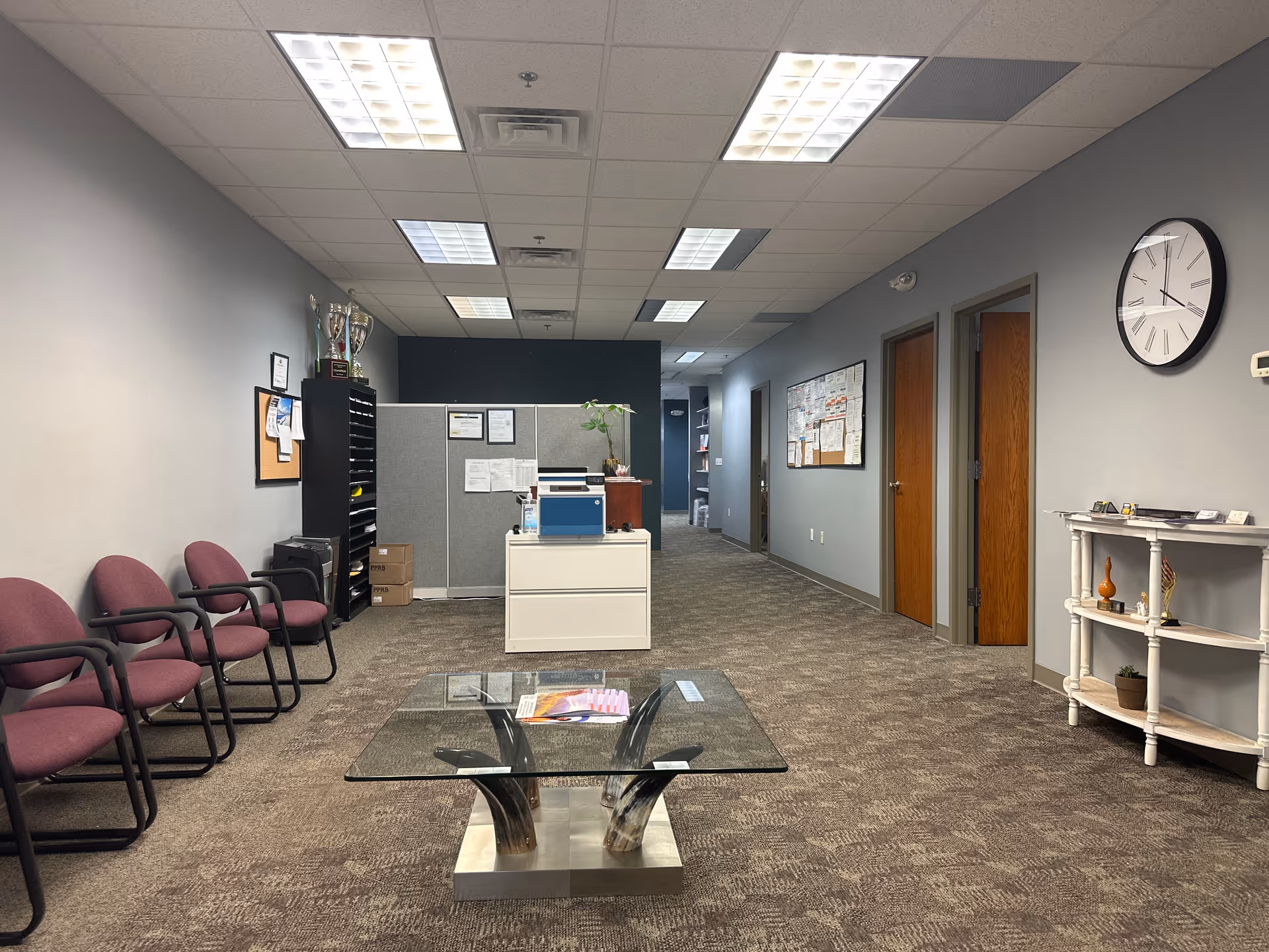 Spacious lobby with burgundy chairs along left, glass coffee table, reception desk, and wall clock.