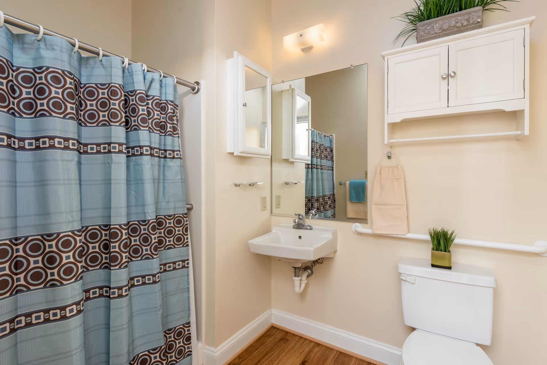 Bathroom in a senior living facility with sink, mirror, toilet, cabinet, and patterned shower curtain.