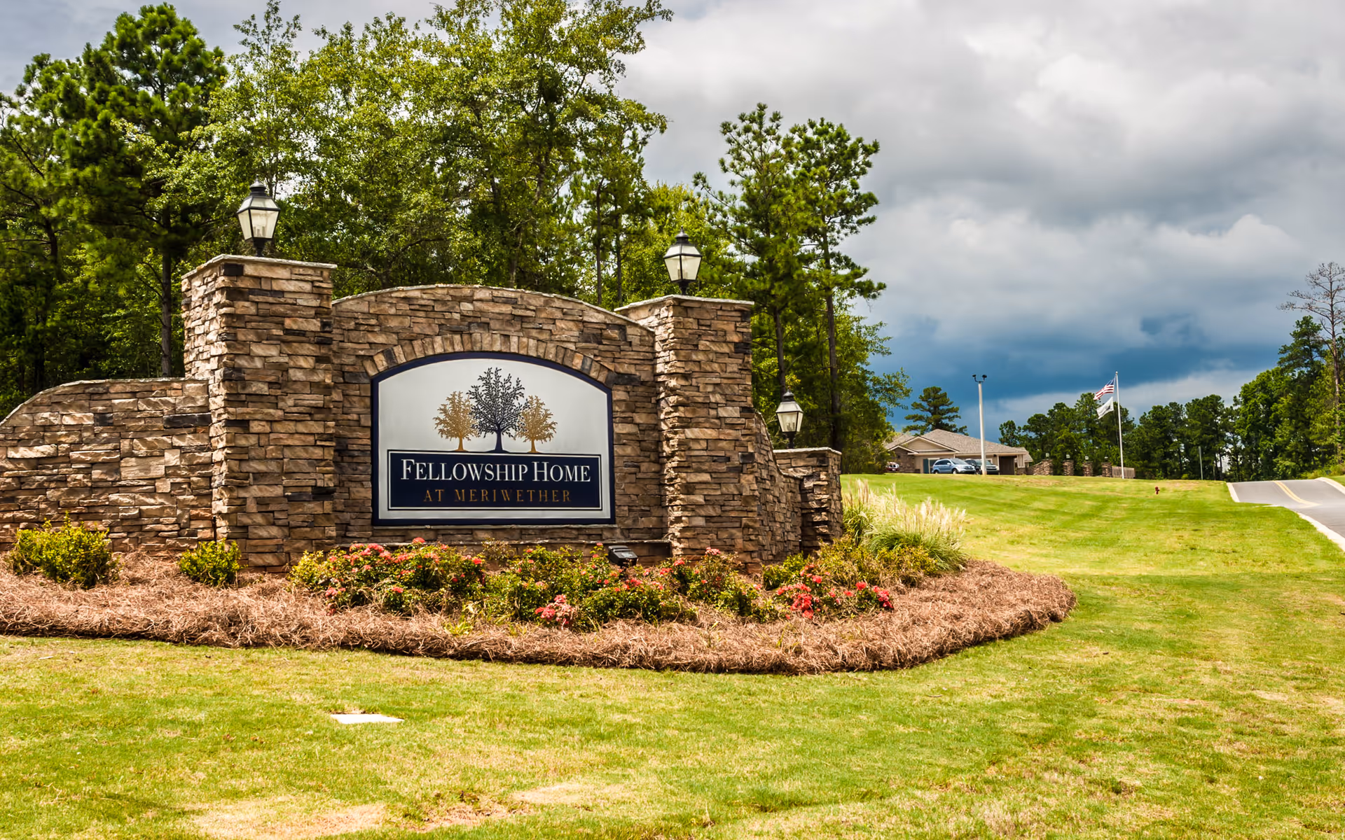Stone entrance sign reading 'Fellowship Home at Meriwether' set in landscaped grass with trees and a cloudy sky.