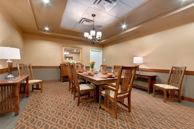 Communal dining room with a wooden table set with place settings, surrounded by wooden chairs, side tables, and warm overhead lighting.