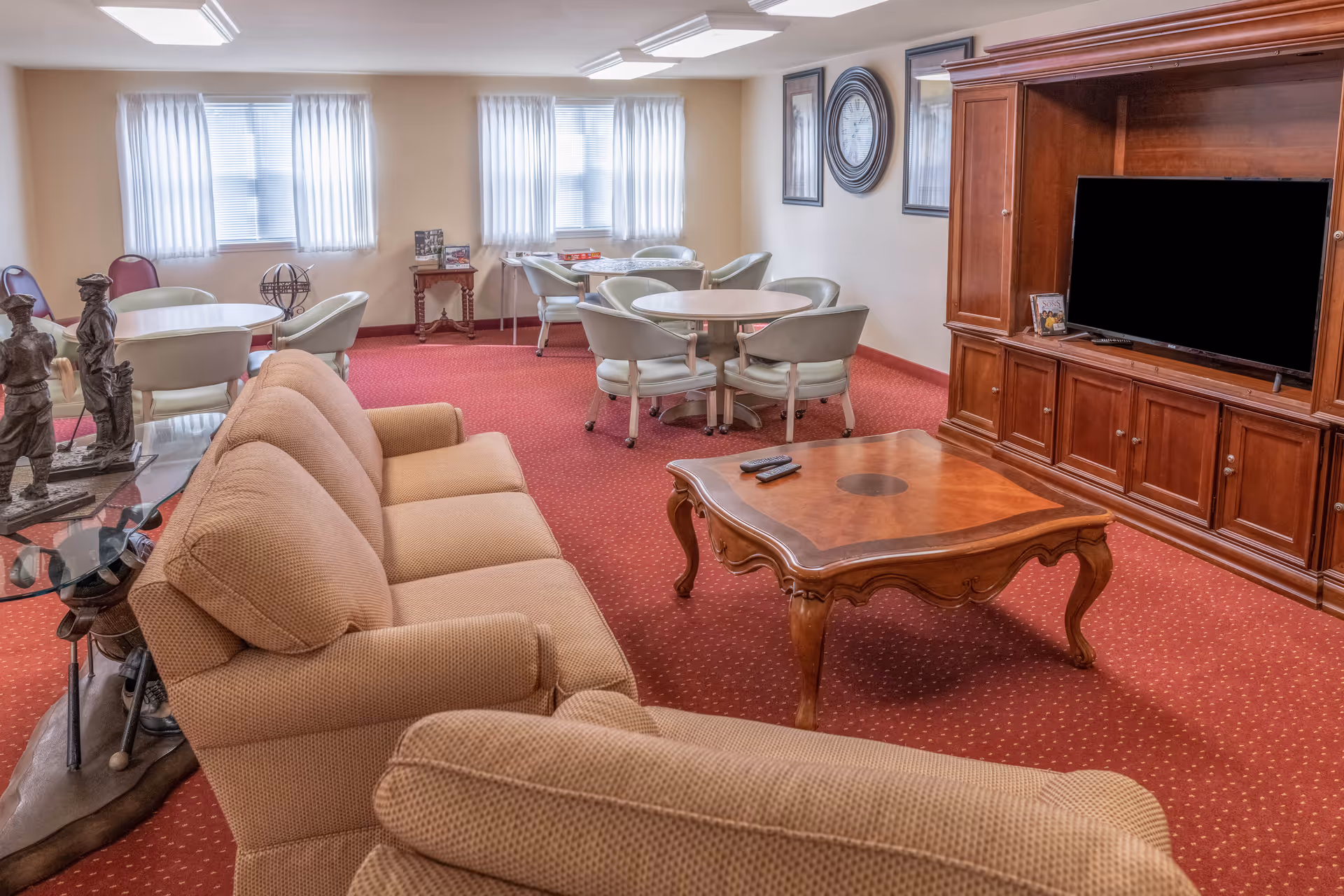 A cozy living room with beige upholstered sofas, a wooden coffee table with two remote controls, and a large wooden entertainment center with a flat-screen TV. The room has red carpet flooring and cream-colored walls with framed artwork and a large clock. In the background, there are round tables with chairs near windows covered with sheer white curtains.