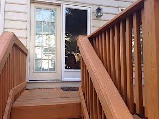 Wooden stairs with railings leading up to a white door with a window and a glass storm door, attached to the exterior of a light-colored building.