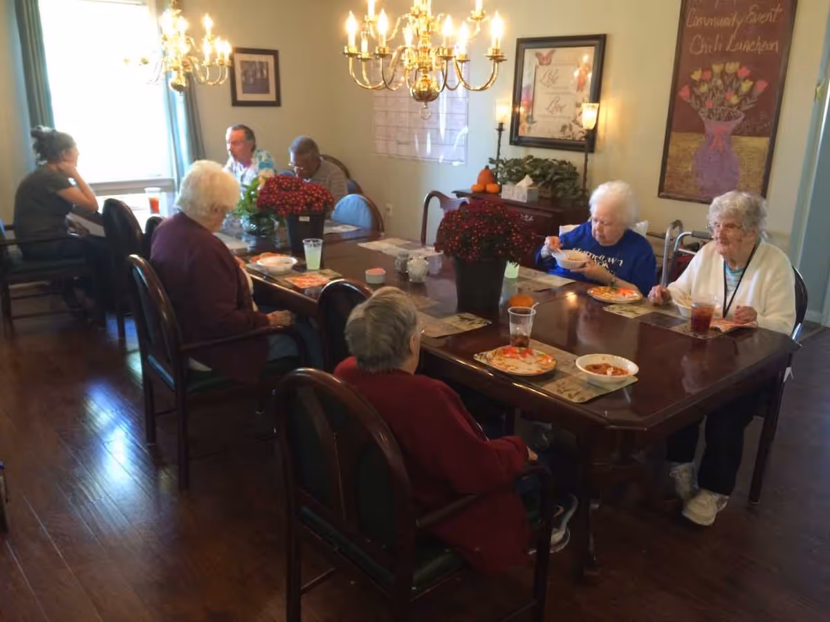 A group of elderly residents seated around a dining table eating and chatting in a communal dining room with chandeliers and wall art.