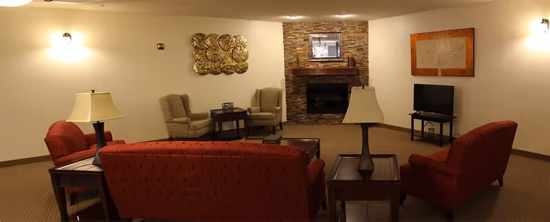 A cozy common room with red sofas, armchairs, side tables, lamps, a TV, and a stone fireplace.