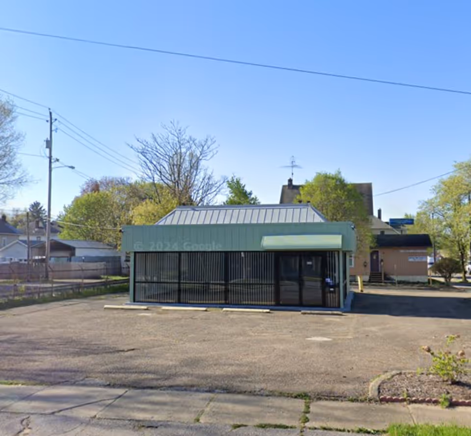 Small green storefront building with glass windows and a parking lot in front.