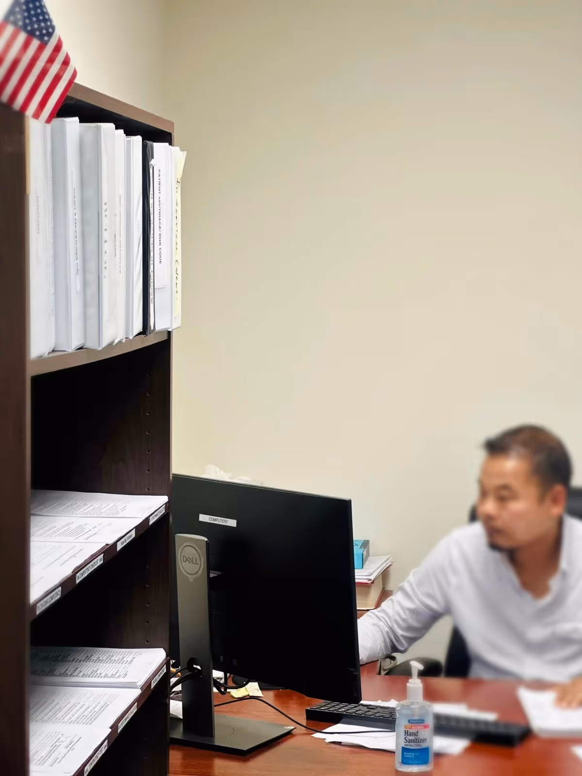 Office scene with a man at a desk, computer, filing shelves, and hand sanitizer.