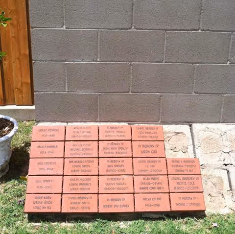 Row of brick memorial plaques on grass beside a gray cinder block exterior wall.