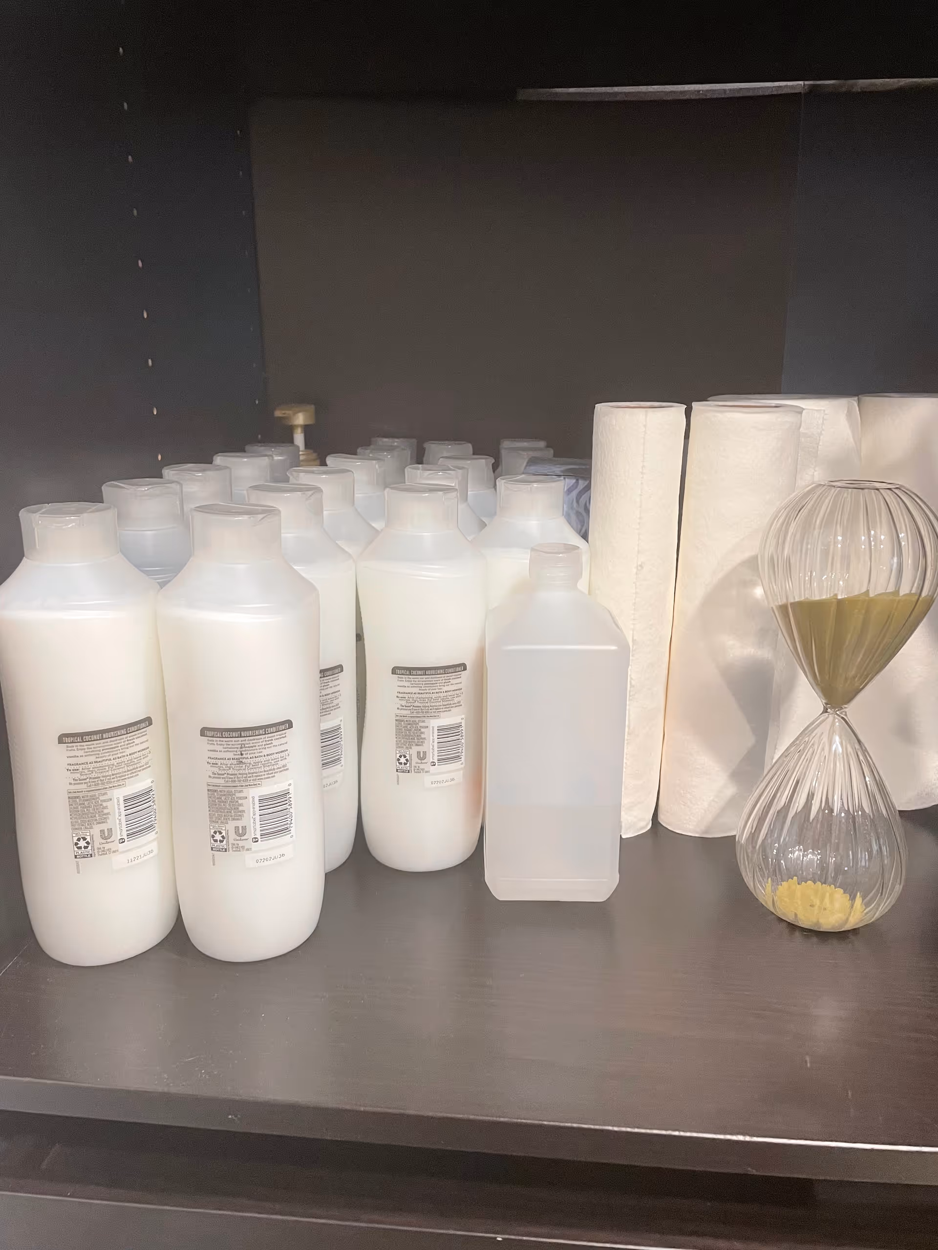 Shelves hold white bottles, a clear jug, paper towels, and an hourglass timer.