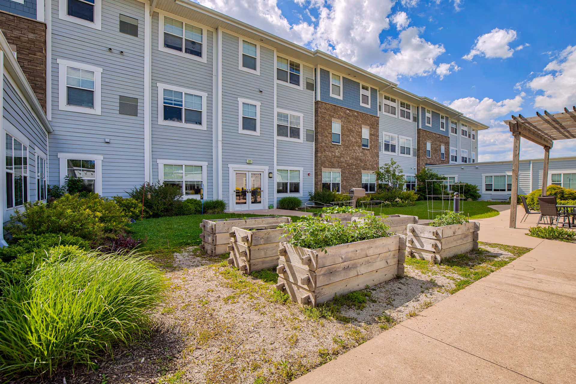 Outdoor garden area at StoryPoint Schererville featuring raised wooden garden beds with plants, surrounded by a paved walkway and green shrubs. The background shows a multi-story building with light blue siding and brick accents under a partly cloudy sky.