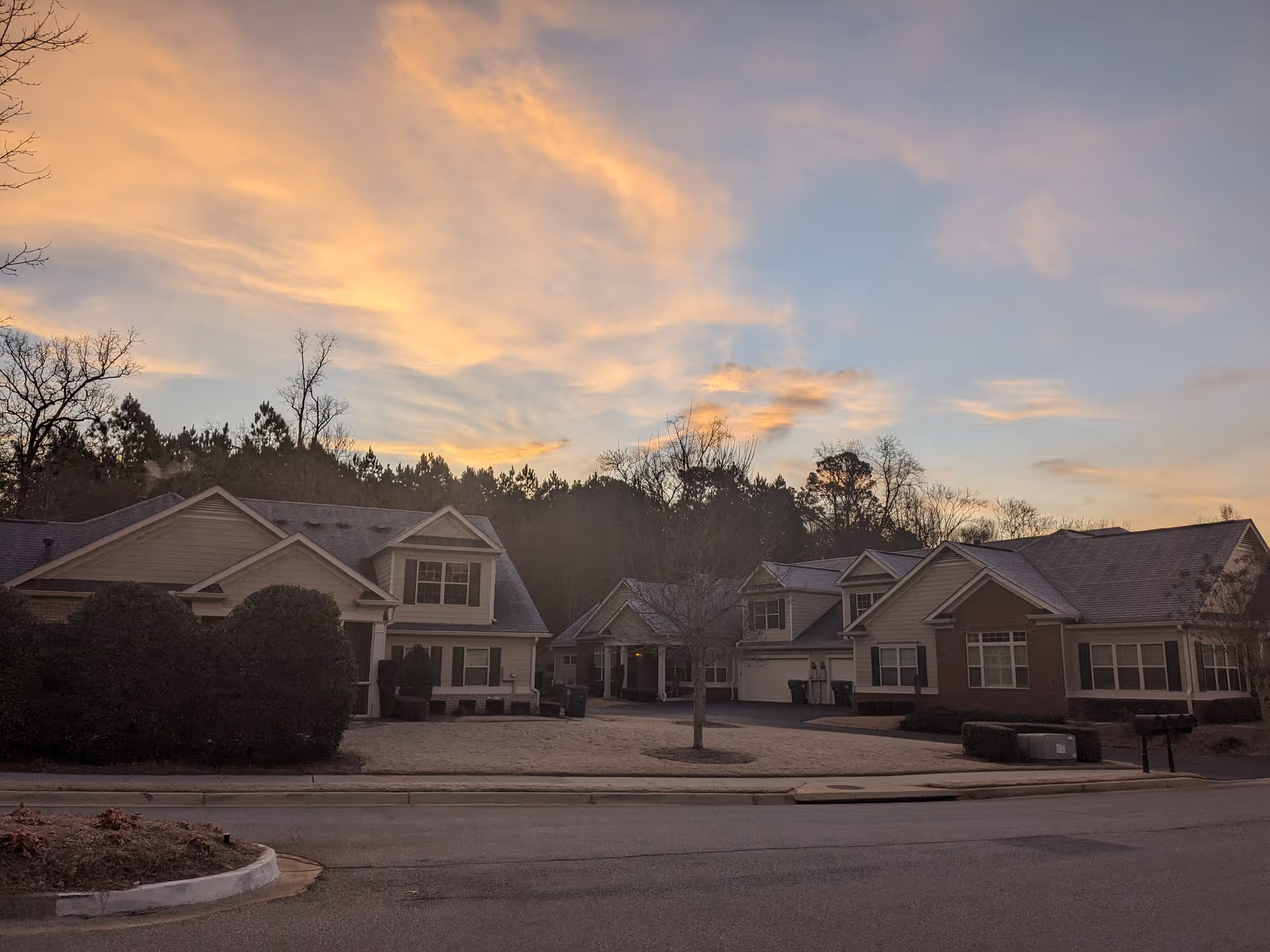 Row of pastel townhomes with driveways, trees, and a sunset sky above.