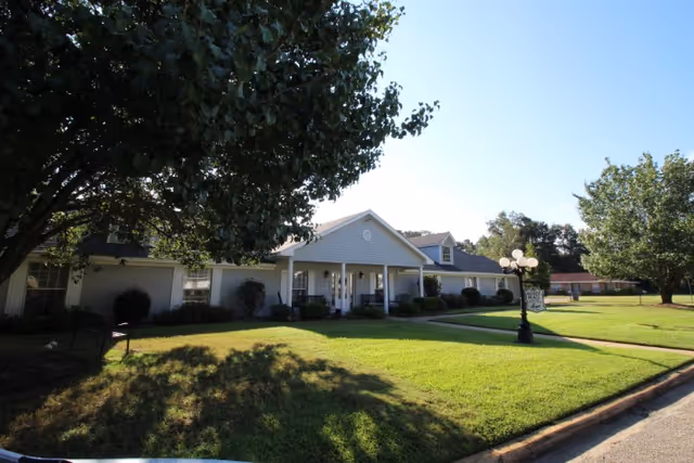 Front exterior of a single-story care building with a covered entrance, manicured lawn, trees, and a decorative lamp post.