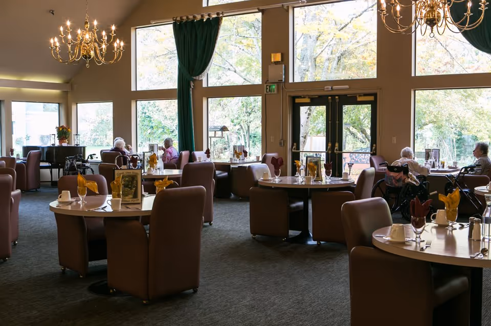 A bright dining room in a senior living facility with round tables, chairs, and residents.