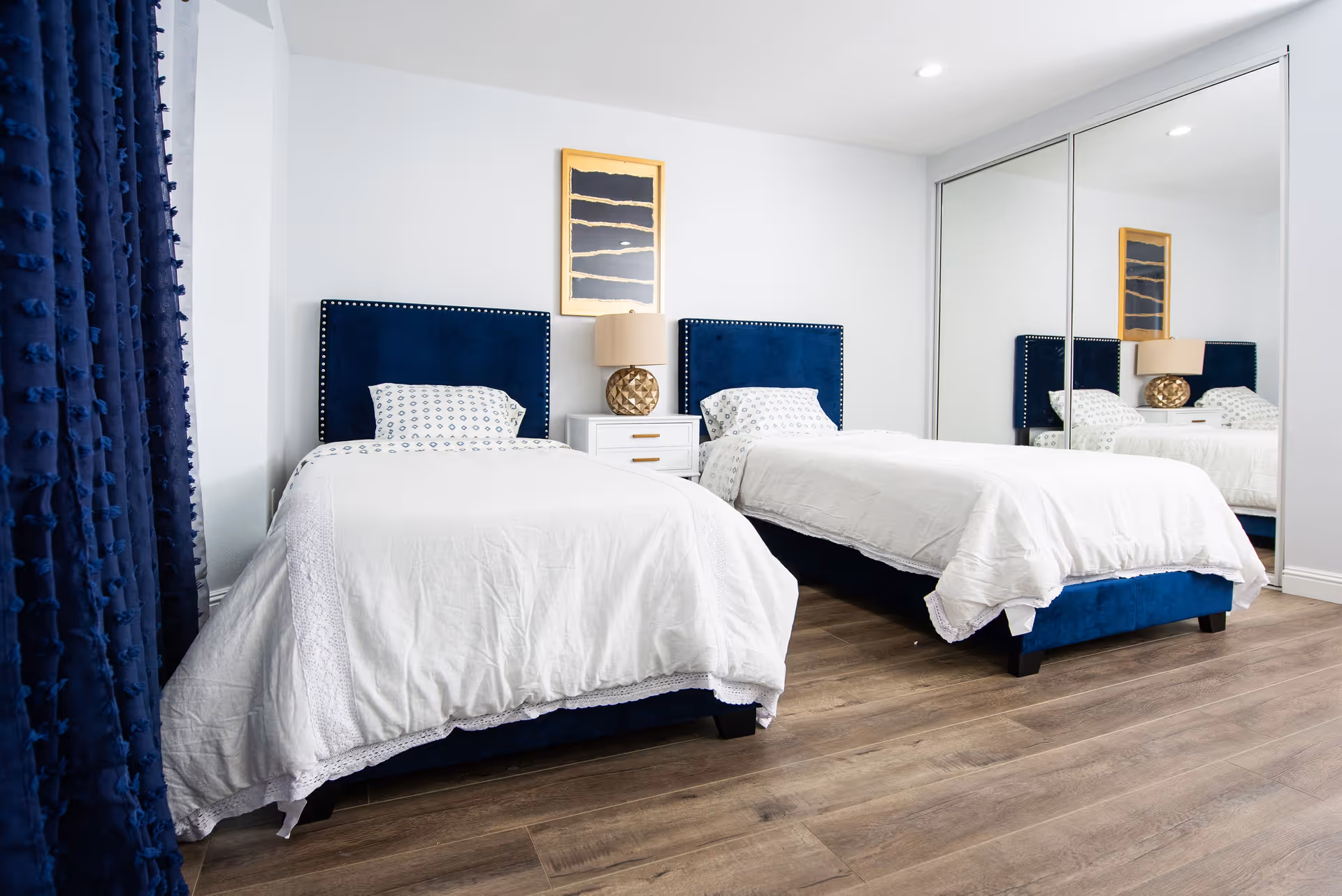 Bright bedroom with two twin beds featuring navy upholstered headboards, white bedding, a nightstand with a lamp, and mirrored closet doors.