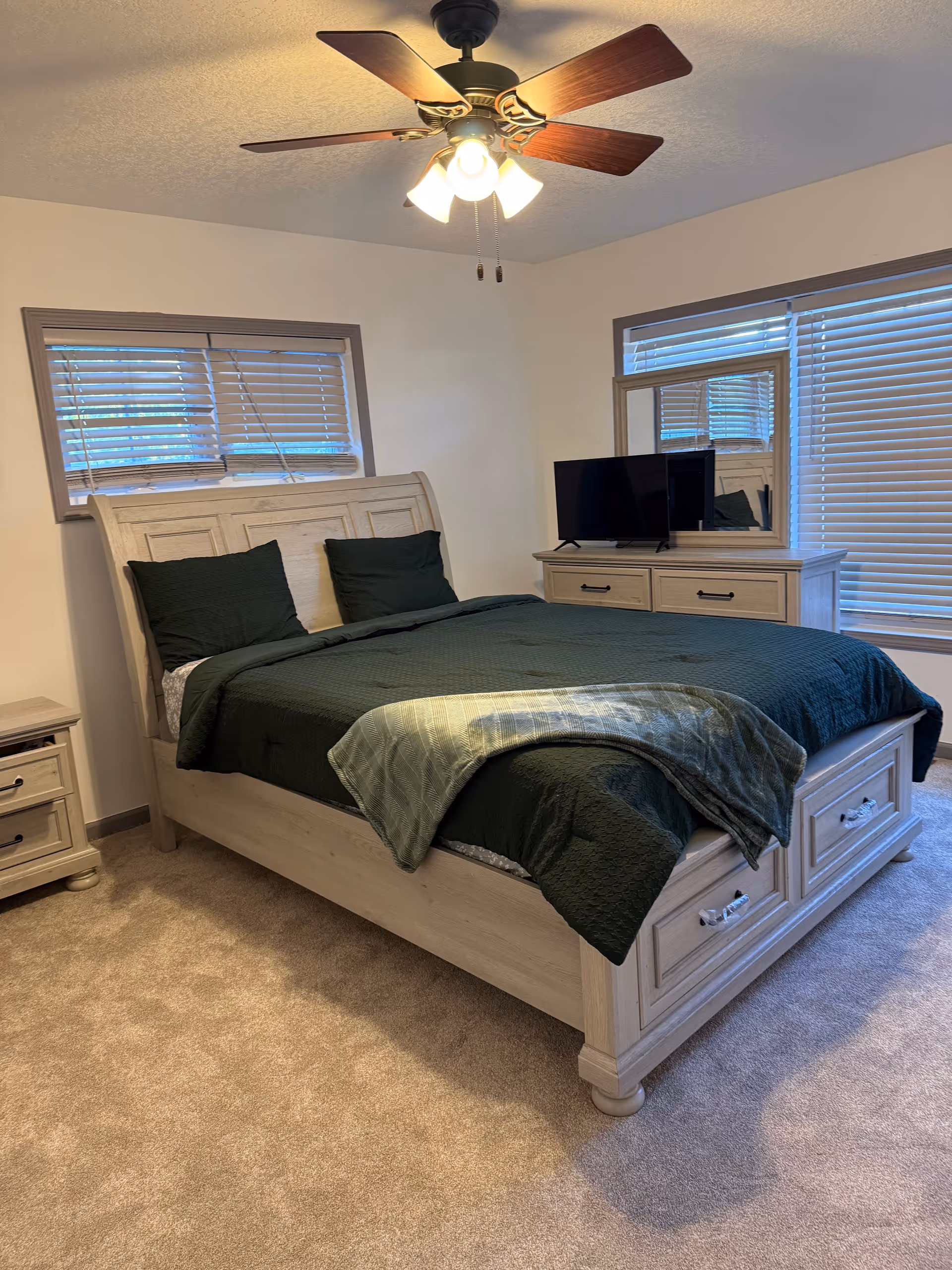 Light wood bedroom with a large bed, dresser, TV, ceiling fan, and blinds.