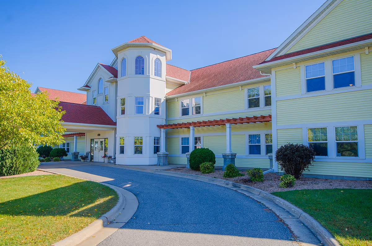Exterior view of a senior living facility named French Meadow Place with light yellow siding, a red roof, and a curved driveway leading to the entrance. The building features multiple windows and a small tower-like structure. There are green bushes and trees around the building under a clear blue sky.
