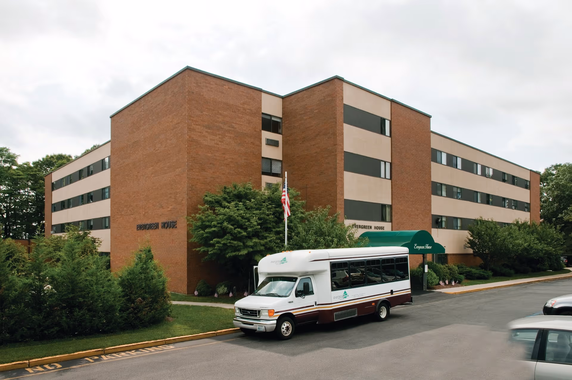 Brick four-story Evergreen House Health Center building with a shuttle bus parked in front and an American flag by the entrance.