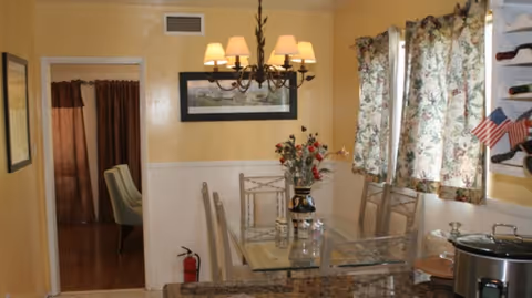 Small dining area with a glass-top table, chandelier, floral curtains and yellow walls.