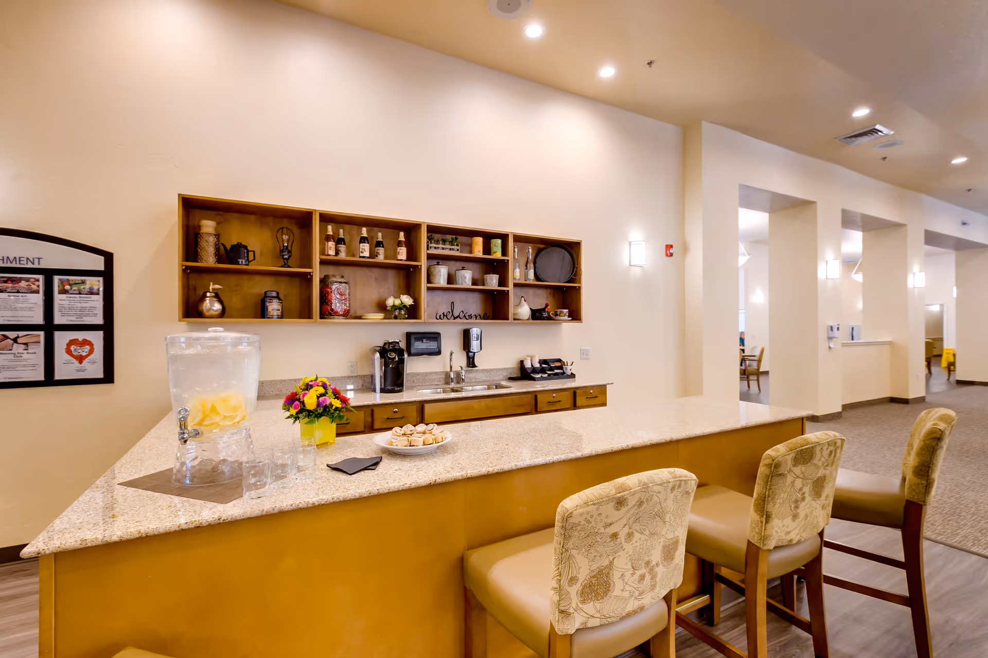 Bright communal kitchenette with a long granite counter, three padded bar stools, a beverage dispenser, coffee station, and open shelving with decor.