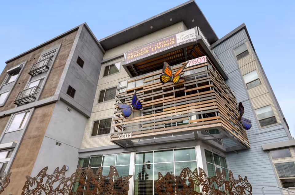Exterior view of a modern multi-story senior living facility with balconies decorated with large butterfly sculptures and a metal butterfly fence in front. The building has a sign that reads 'Modern Urban Senior Living Assisted Living & Memory Care' and the address number 7601 is visible.