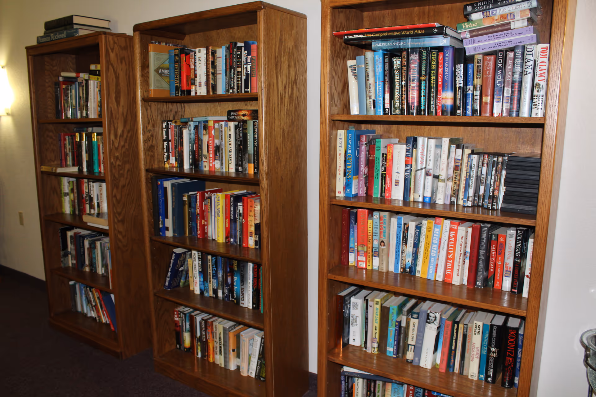 Three wooden bookshelves filled with a variety of books in a well-lit indoor space with a carpeted floor and a wall-mounted light fixture.