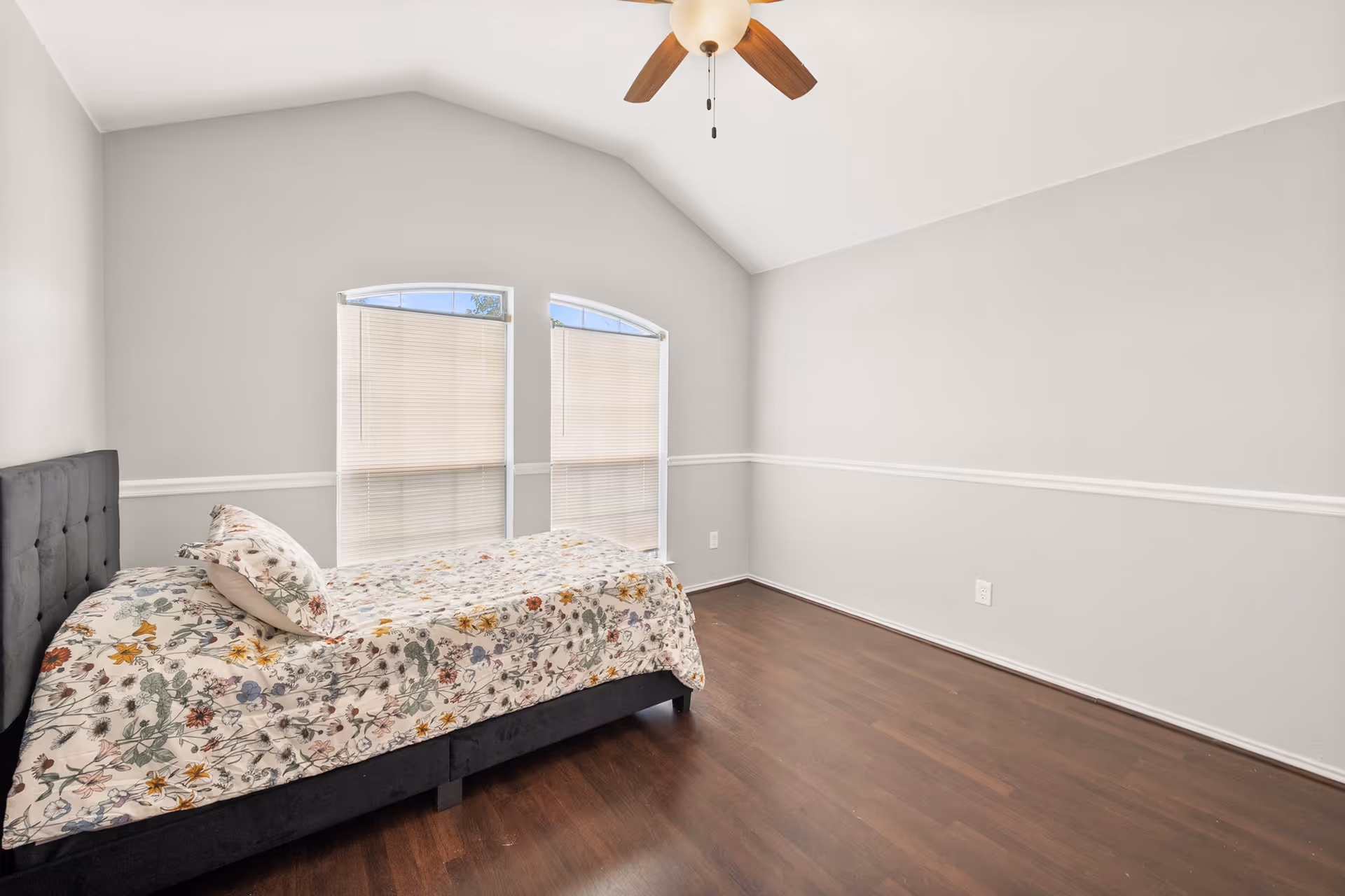 A sparsely furnished bedroom with a bed, windows, and ceiling fan.