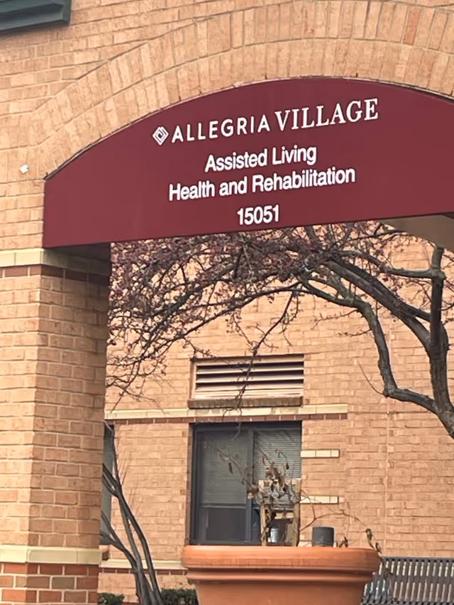 Brick building exterior with a maroon Allegria Village sign for assisted living.