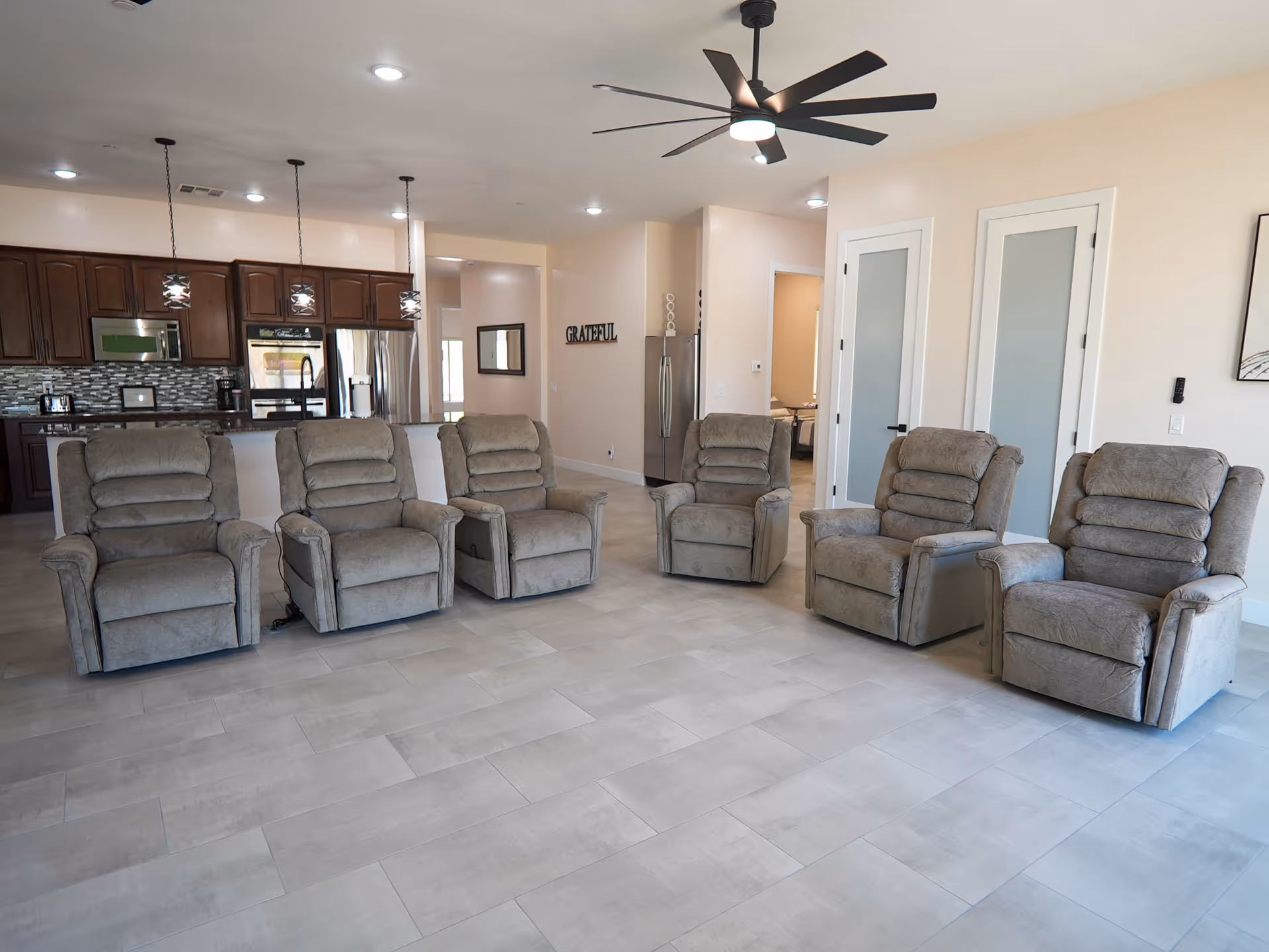 Open communal living area with six gray recliner chairs, kitchen, and modern ceiling fan.
