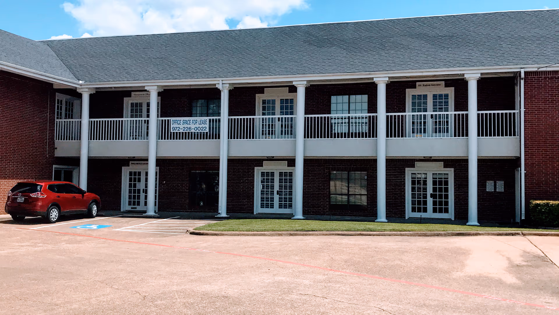 Exterior view of brick two-story building with white columns and a parked red car.