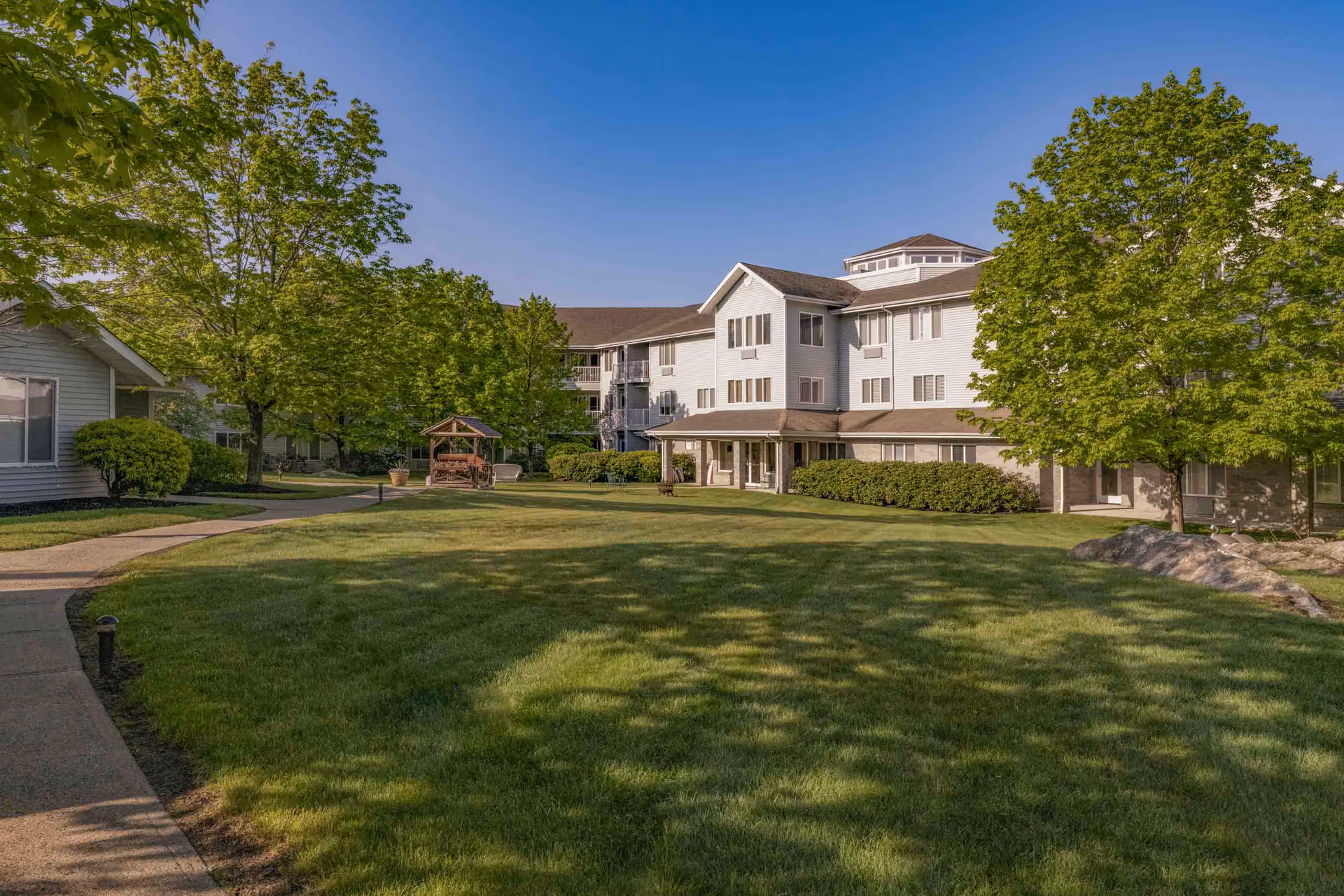 Sunlit courtyard with a grassy lawn, walkway, trees and a gazebo in front of a multi-story senior living building.