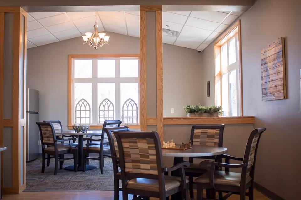 Communal dining area with round tables, upholstered chairs, a chess set, and large windows letting in natural light.