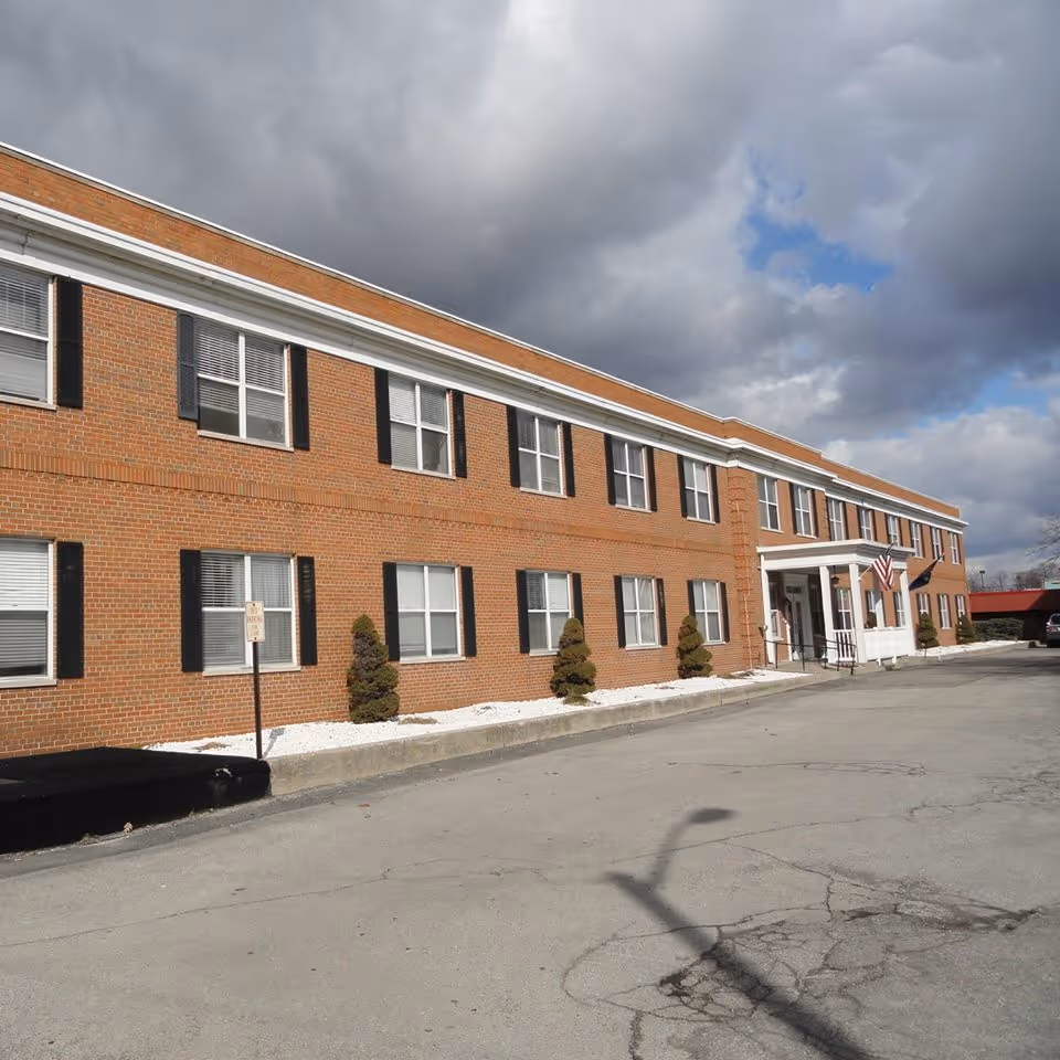 Front exterior of a two-story brick senior living building with rows of windows and an American flag at the entrance under a cloudy sky.