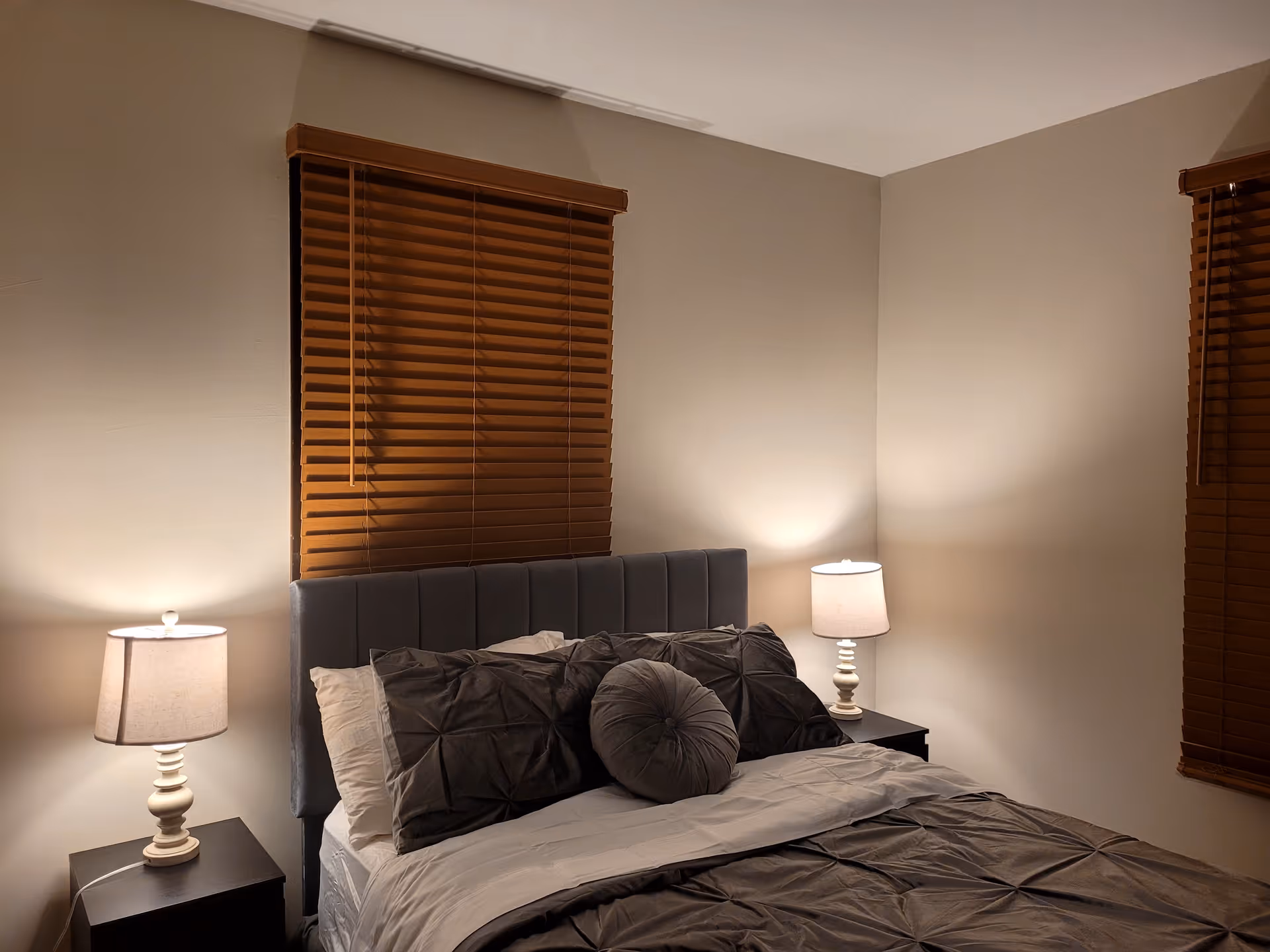 A neatly made bedroom with a padded headboard, two nightstands, lamps, and wooden blinds.